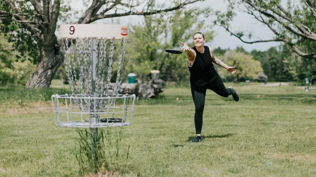 Osoba rzuca frisbee w stronę kosza do disc golfa w parku.