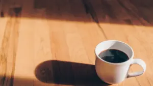 Biały kubek z czarną kawą na drewnianym stole w promieniach popołudniowego słońca.