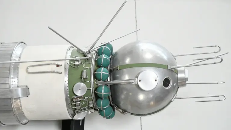 Model metalowego satelity kosmicznego z okrągłą sekcją i wystającymi antenami na jasnym tle.